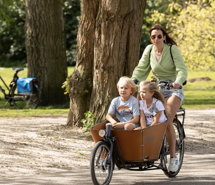 Young woman cycles with her cargo bike with children through the Vondelpark in Amsterdam